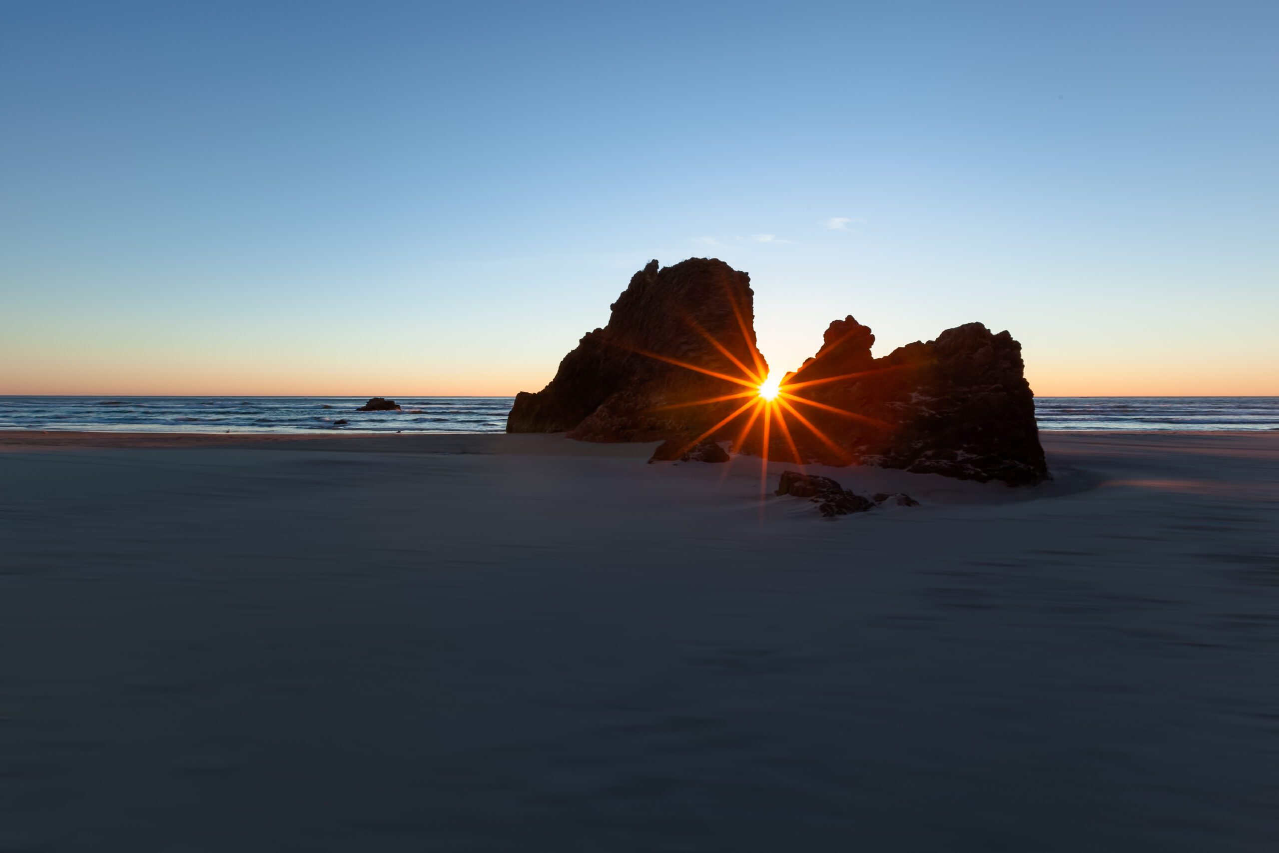 Experience the serene closure of day with this mesmerizing capture, where the Pacific kisses the sun goodbye through dramatic rocky silhouette .