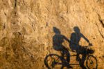 Tranquil synergy of light and shadow, as cyclists cast elongated figures on a sunlit rock wall, epitomizing Vancouver’s vibrant seawall in Stanley Park.