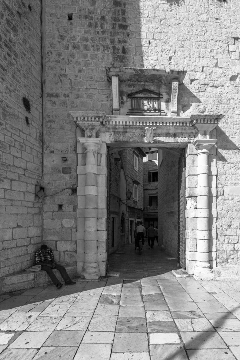 A grayscale image capturing the essence of historical Trogir, Croatia, focusing on an architectural doorway, with a local resident immersed in a moment of solitude, complemented by the cobbled street and distant human elements, symbolizing the blend of everyday life with the town's ancient allure.