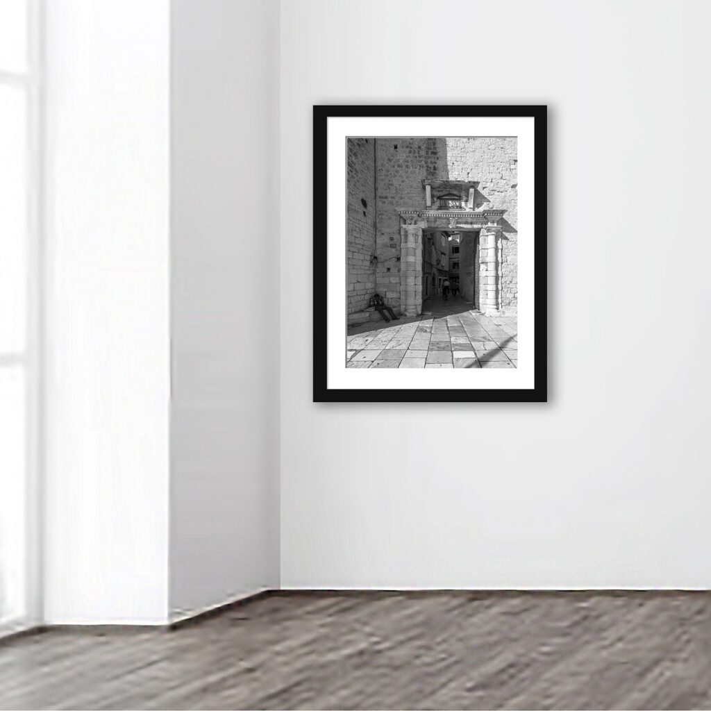 A grayscale image capturing the essence of historical Trogir, Croatia, focusing on an architectural doorway, with a local resident immersed in a moment of solitude, complemented by the cobbled street and distant human elements, symbolizing the blend of everyday life with the town's ancient allure.