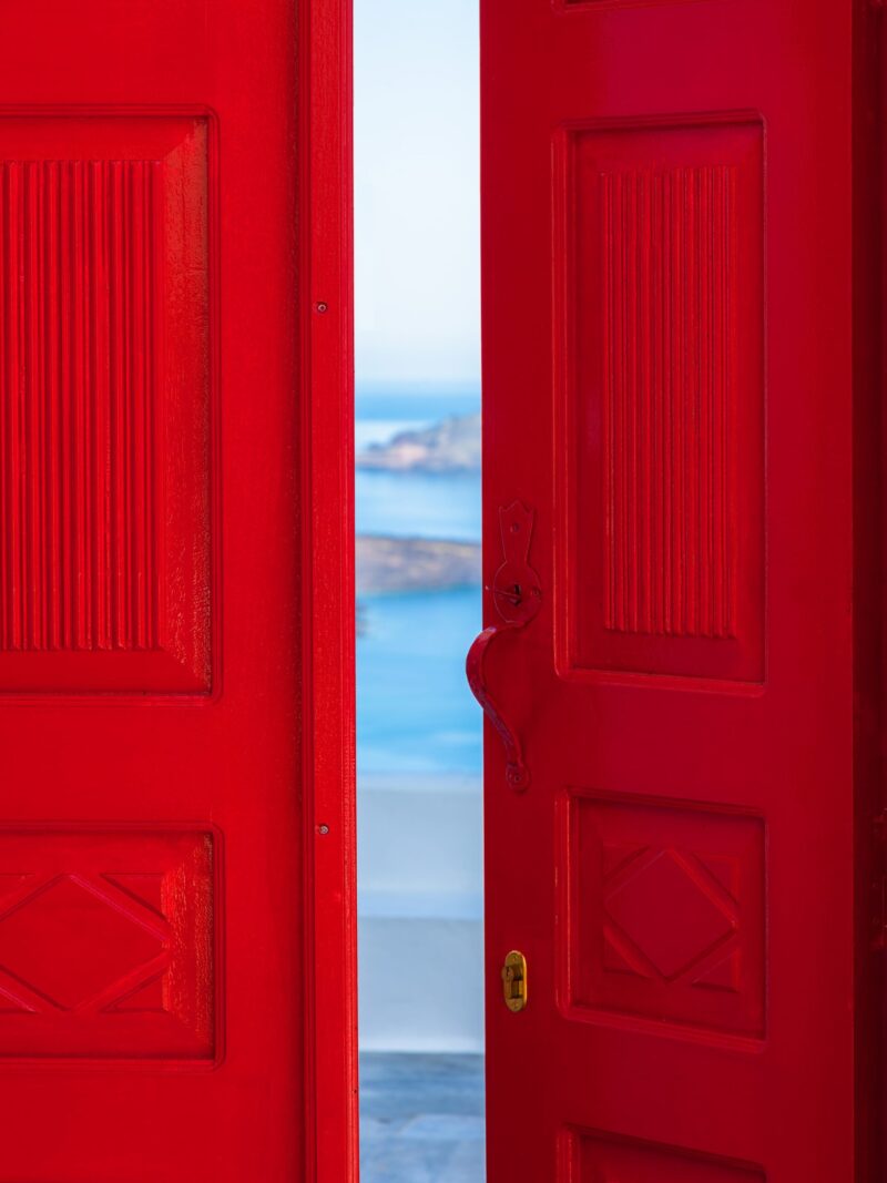 Bright red doorway opening to a view of the Santorini caldera with blue waters below.